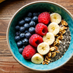 A vibrant smoothie bowl topped with fresh berries, granola, chia seeds, and sliced banana, arranged in an artistic pattern. The smoothie bowl itself should have a green hue, suggesting spinach or kale, and be served in a rustic ceramic bowl. Sunlight streams in from a nearby window, highlighting the textures and colors.